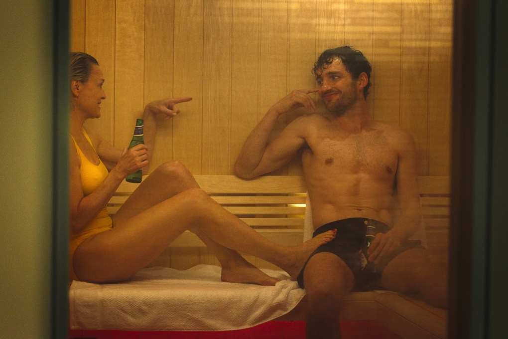 Robin Wright, wearing a yellow swimsuit, and Laurie Davidson, in dark shorts, sitting in a wooden sauna with bottles of beer. Wright is pointing at Davidson, who is smiling.