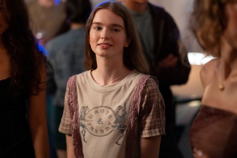 A girl in a cream Haute & Freddy T-shirt stands smiling at a party in Prime Video's Carrie.